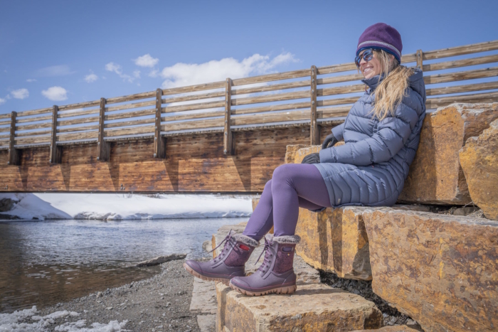 Woman wearing Bogs Arcata Faded winter boots sitting on stone steps beside a snowy river