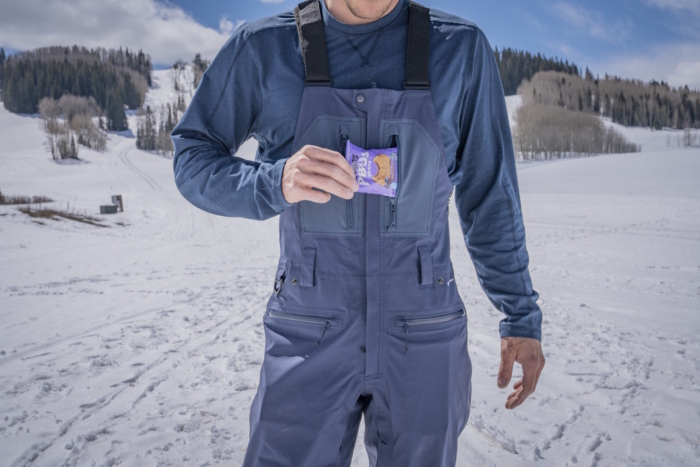 A person wearing a Flylow Stash Bib pulls a purple snack bar out of the chest pocket