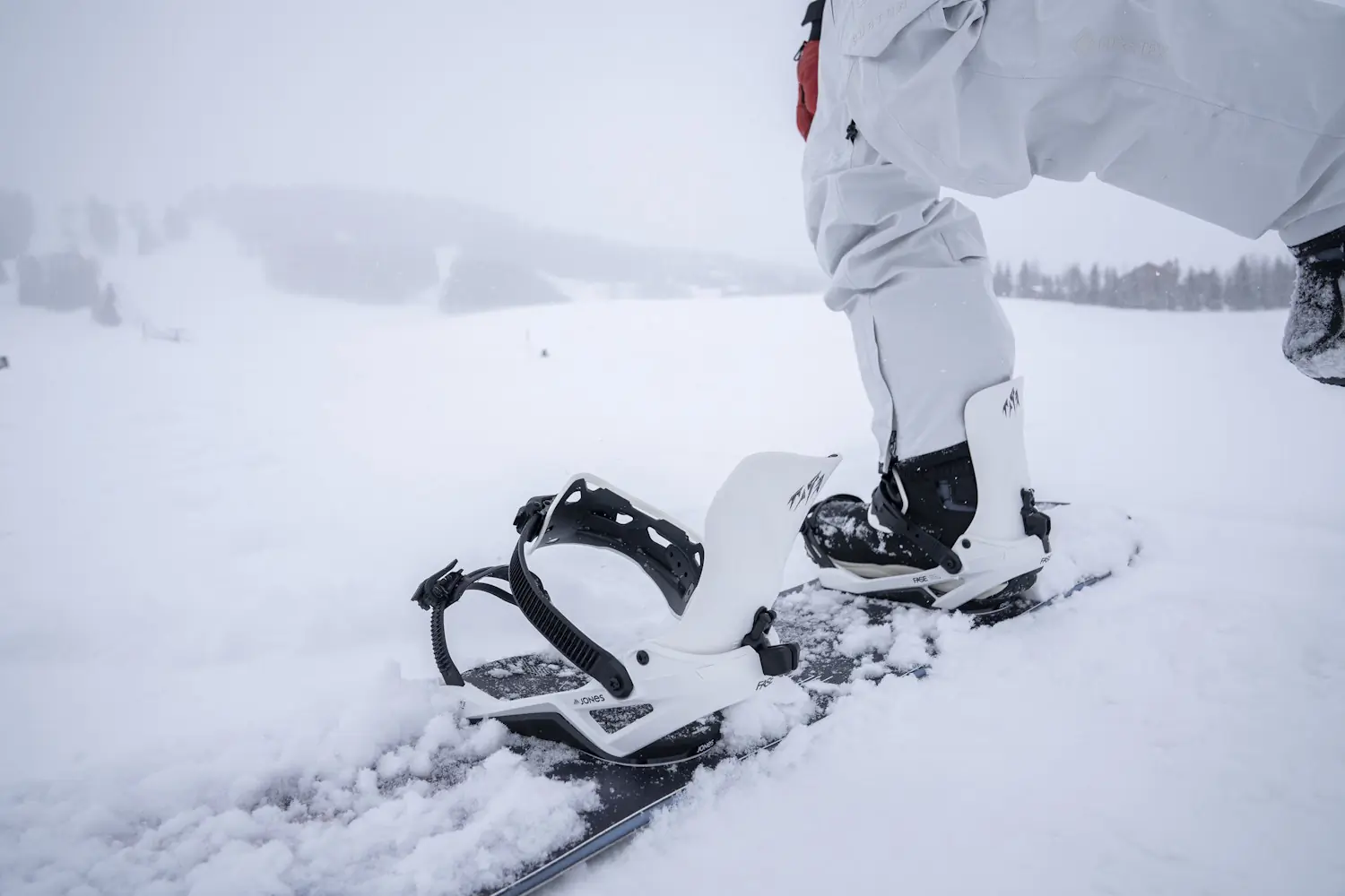 Snowboarder kneeling on a snowy slope with one boot secured in a Jones Mercury FASE binding