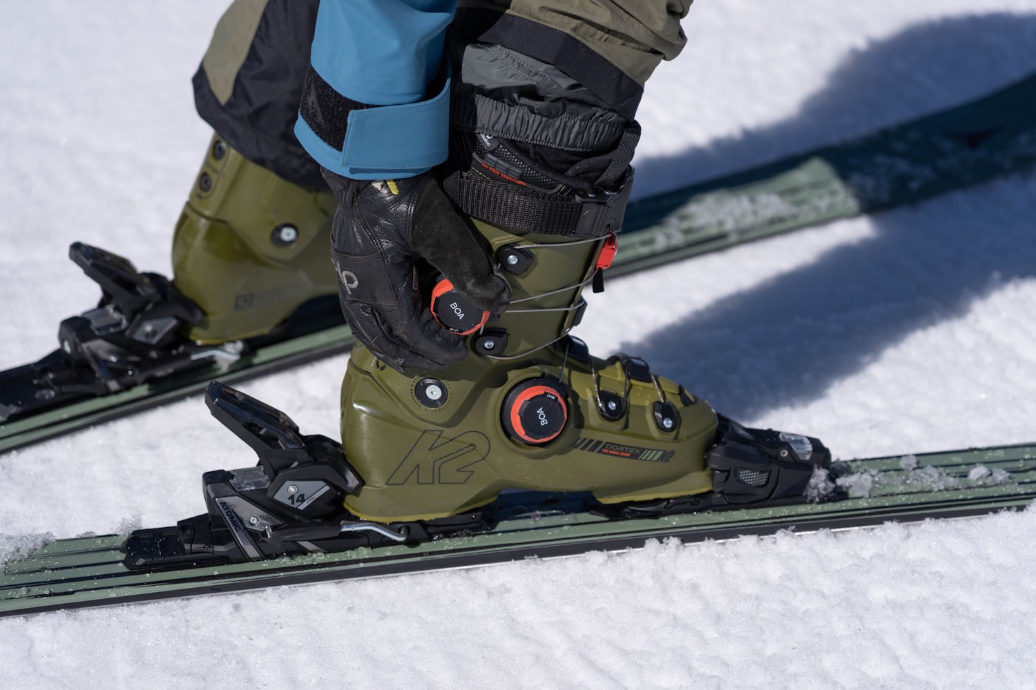 Skier adjusting olive green K2 Cortex 130 Zonal BOA ski boot with dual BOA dials on snowy slope