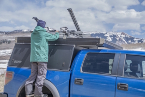 A person loads snowboards onto a Kuat Grip 6 Ski/Snowboard Rack mounted on a blue truck