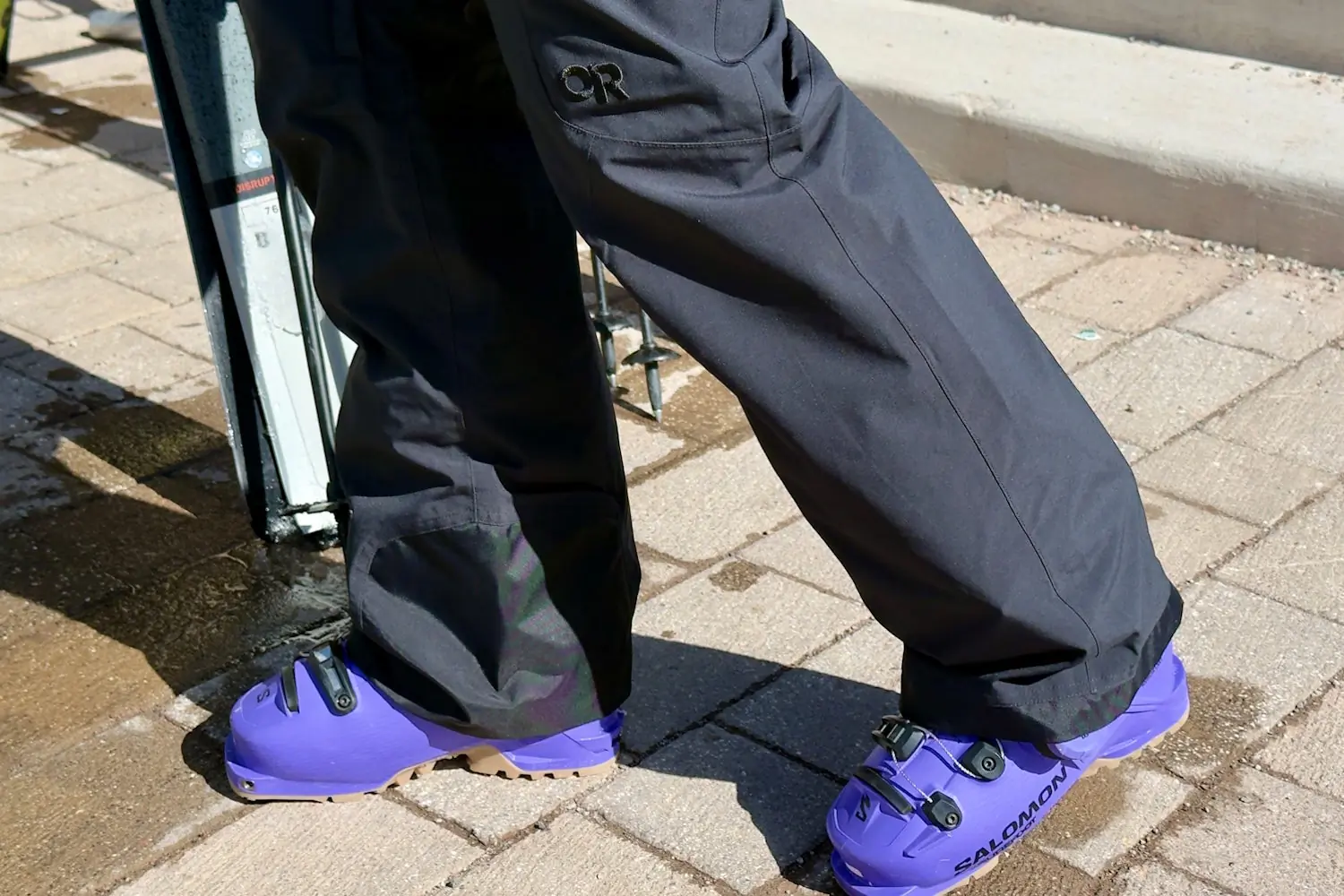 Person in OR Snowcrew pants and purple Salomon ski boots near skis.