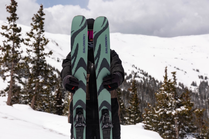 Person holding a pair of Salomon QST 94 Skis with a snowy mountain backdrop.