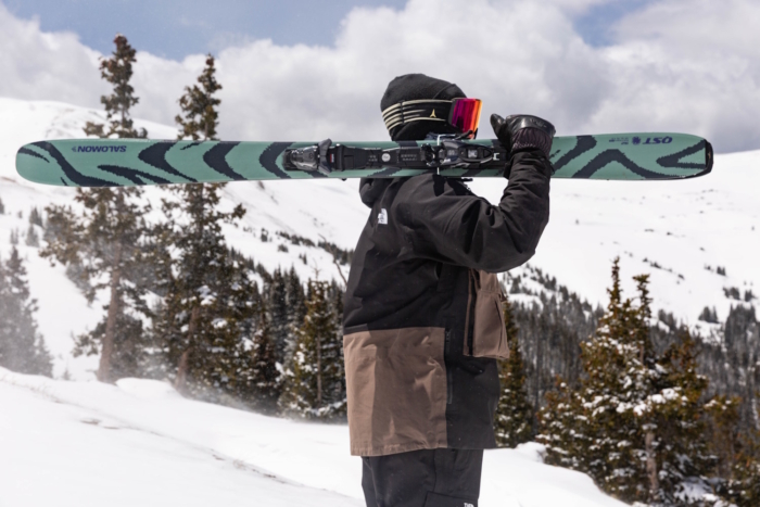 Skier carrying Salomon QST 94 skis in a snowy mountain landscape.