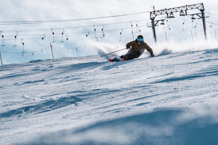 Skier in a brown jacket carving through fresh snow on a sunny slope beneath a ski lift.