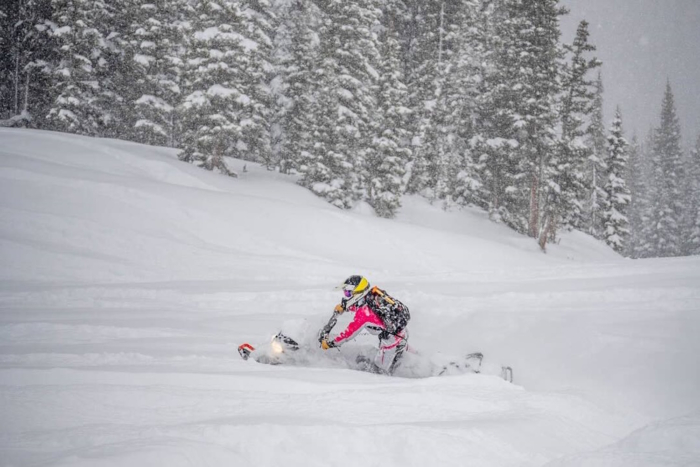 Snowmobiler in pink gear rides through deep powder during snowfall in a forested mountain area.