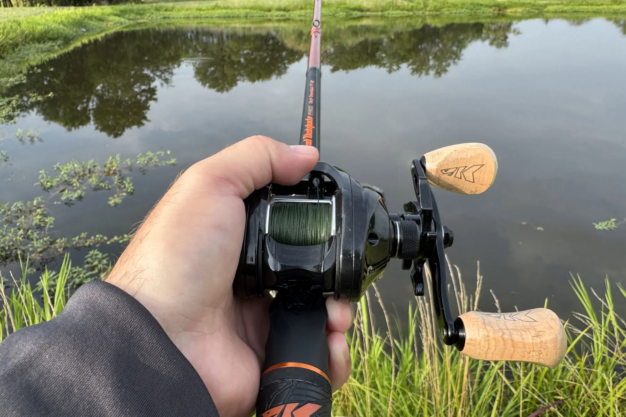 A person holding a baitcasting fishing rod and reel, aimed toward a calm pond