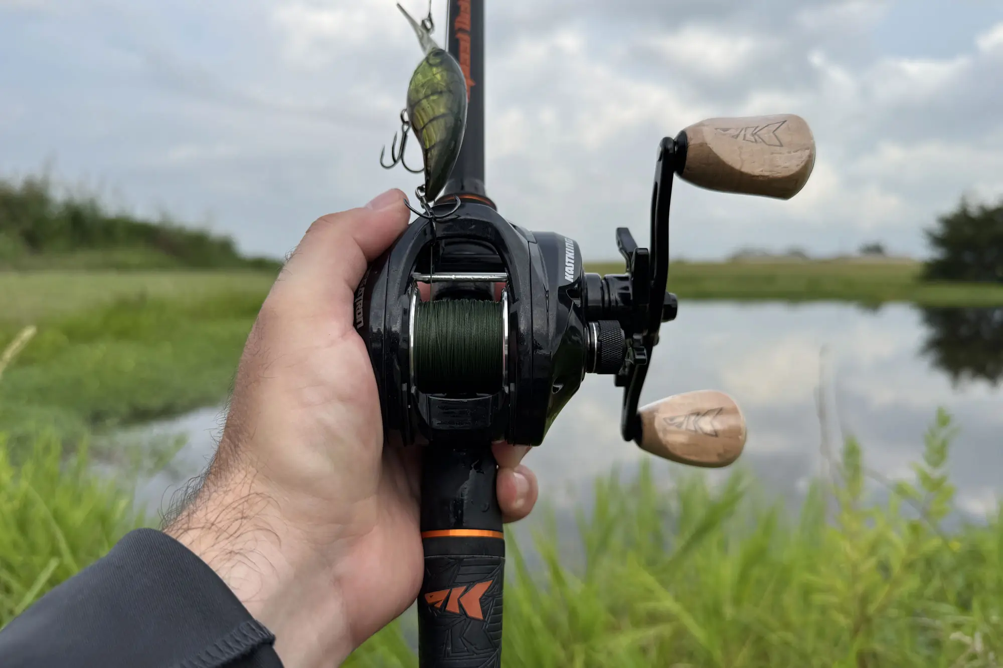 Hand holding baitcasting reel with crankbait by a lake