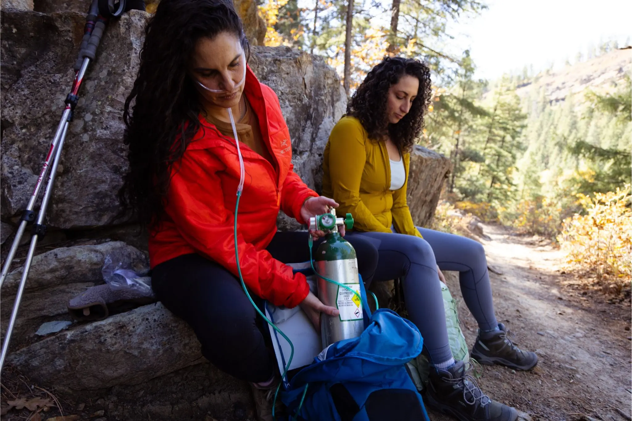 Two people sit side by side on a trail. One wearing a cannula loads a tank of supplemental oxygen into a bag.