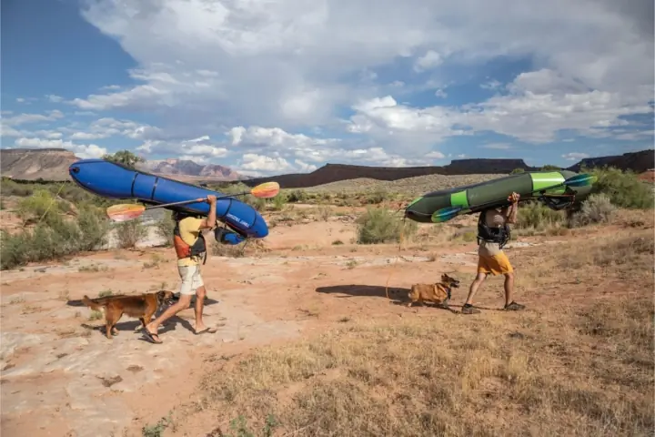 Two people carry canoes above their heads in a desert like landscape