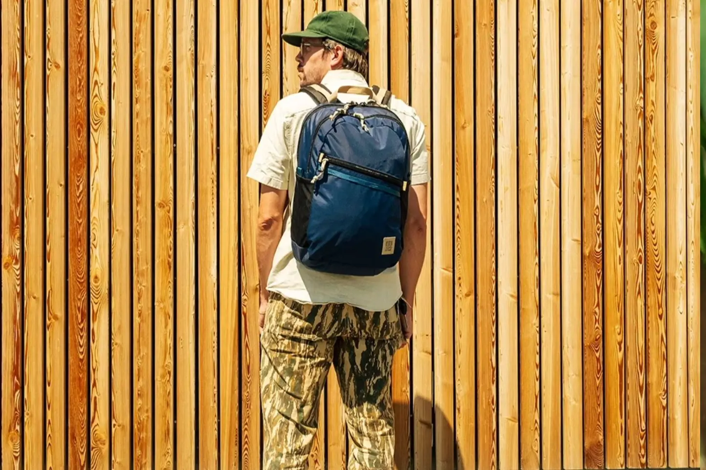 Man in green hat, white shirt, and camo pants wears a blue backpack with back to the camera, in front of a wood paneled wall