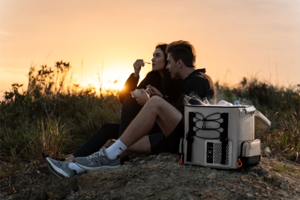 Two people sit on trail eating with backpack fridge in foreground and sunset in background 