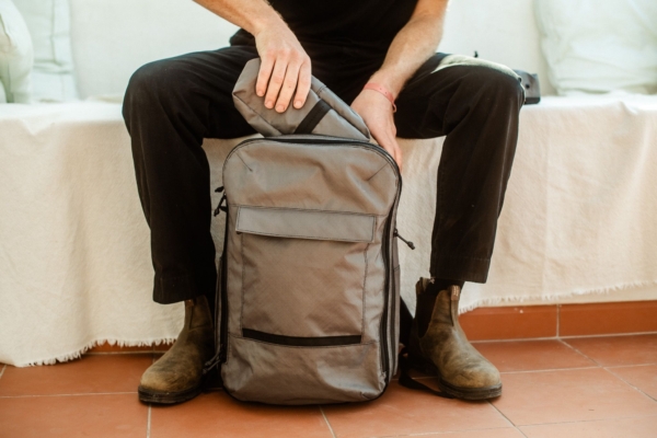 Person in black pants and boots sits and loads a small bag into a larger grey backpack 
