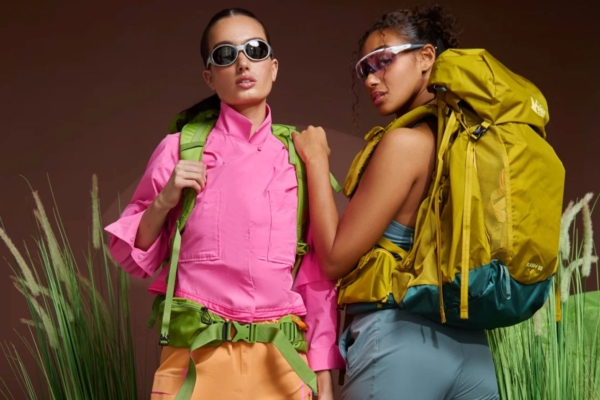 Two women in front of grass and brown background, one wears pink jacket and orange pants, the other has blue top and pants with yellow backpack 