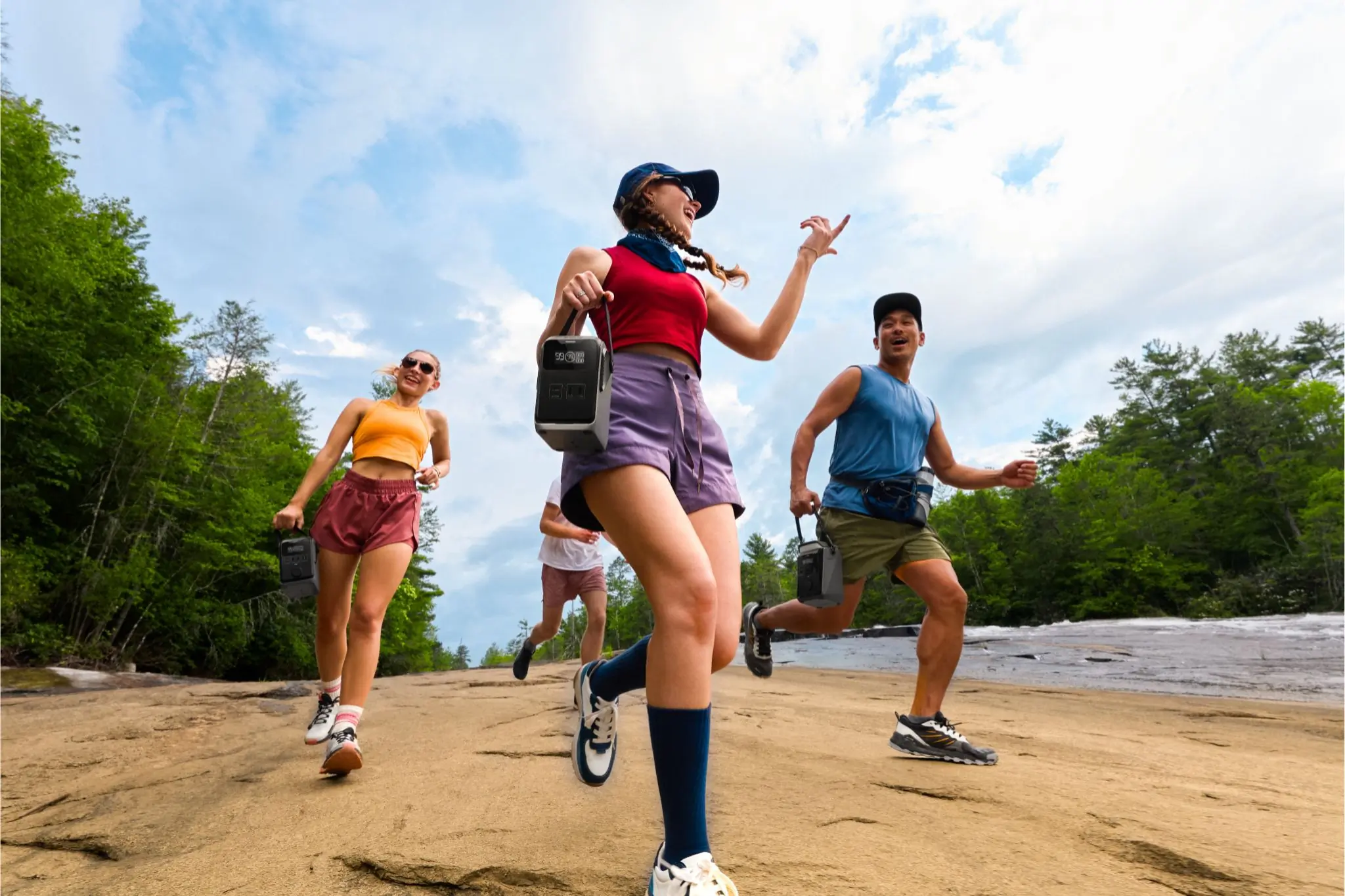 Four hikers carrying power stations walk along a trail with river to the right and trees in the background