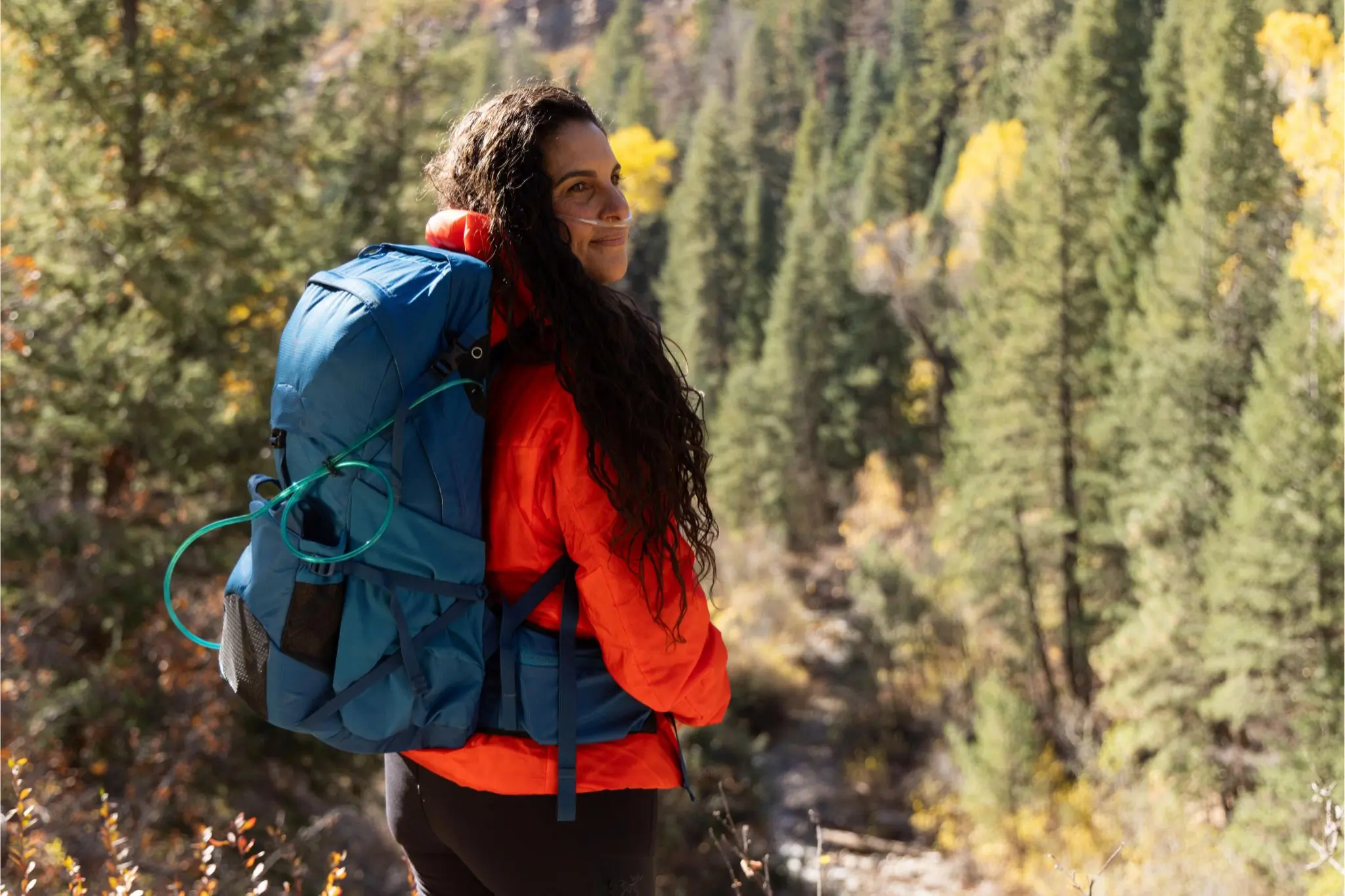 Person in red jacket stands with forest in the background wearing a blue backpack