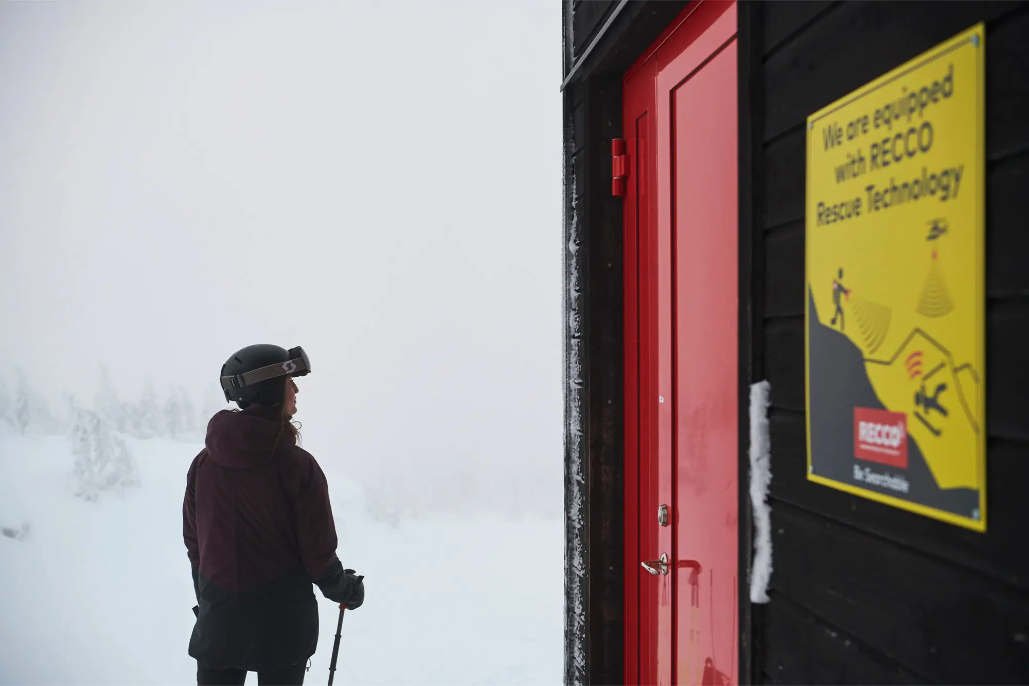 A skier stands by a cabin marked with RECCO rescue signage