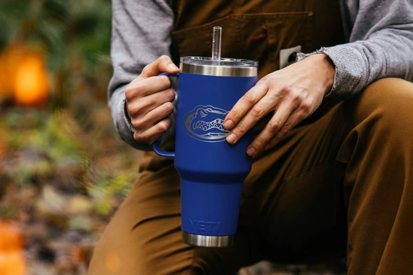 Person in gray top and brown overalls holds large blue mug with Florida Gators logo by a campfire