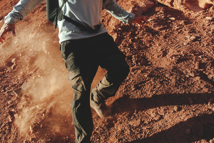 A young male scrambles down a dusty hill of red dirt wearing Columbia ROC pants, demonstrating the durability and ruggedness of the pants.