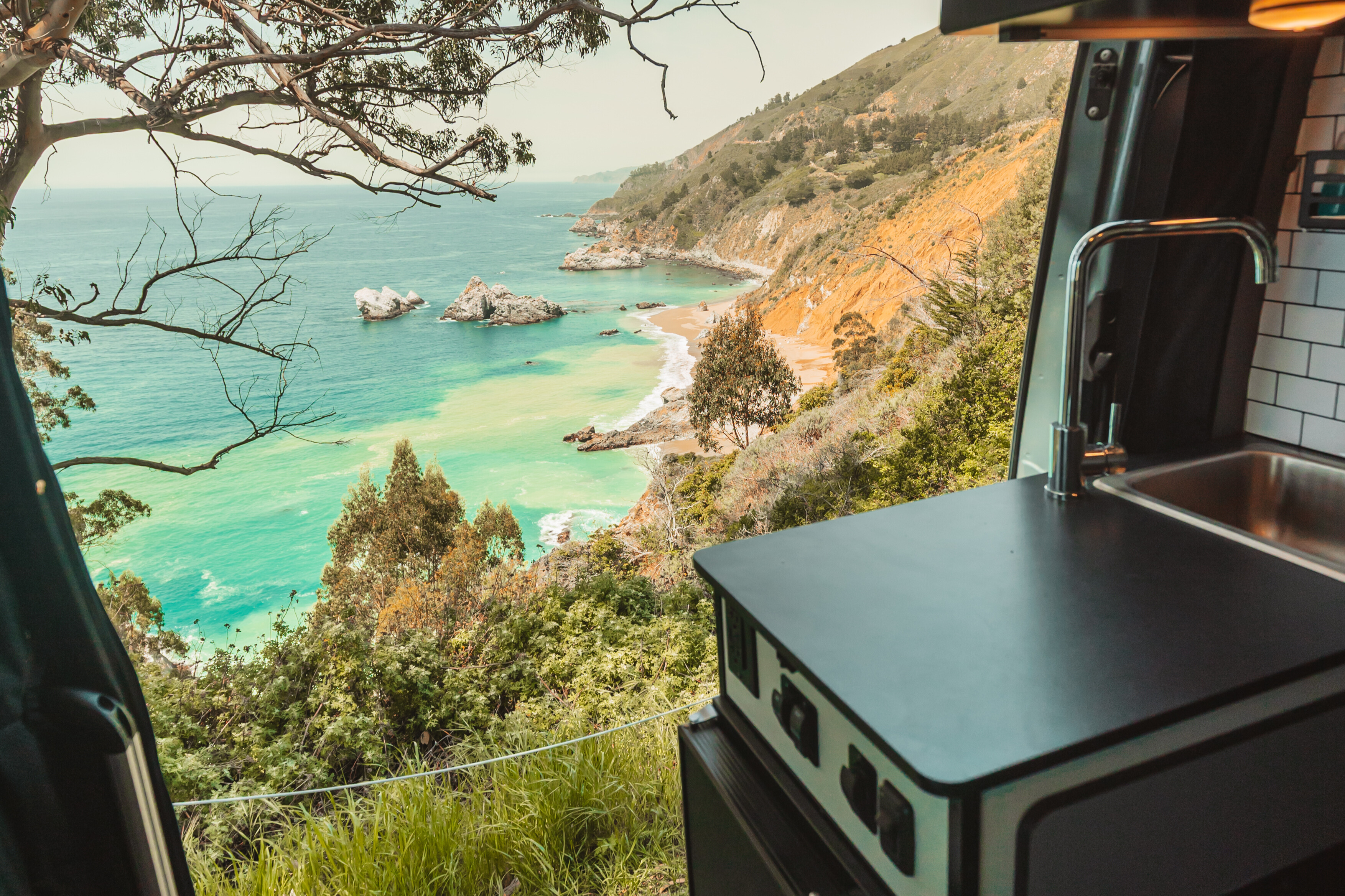 A view of a ocean cliff side overlooking aquamarine waters through the kitchen door of a camper van.