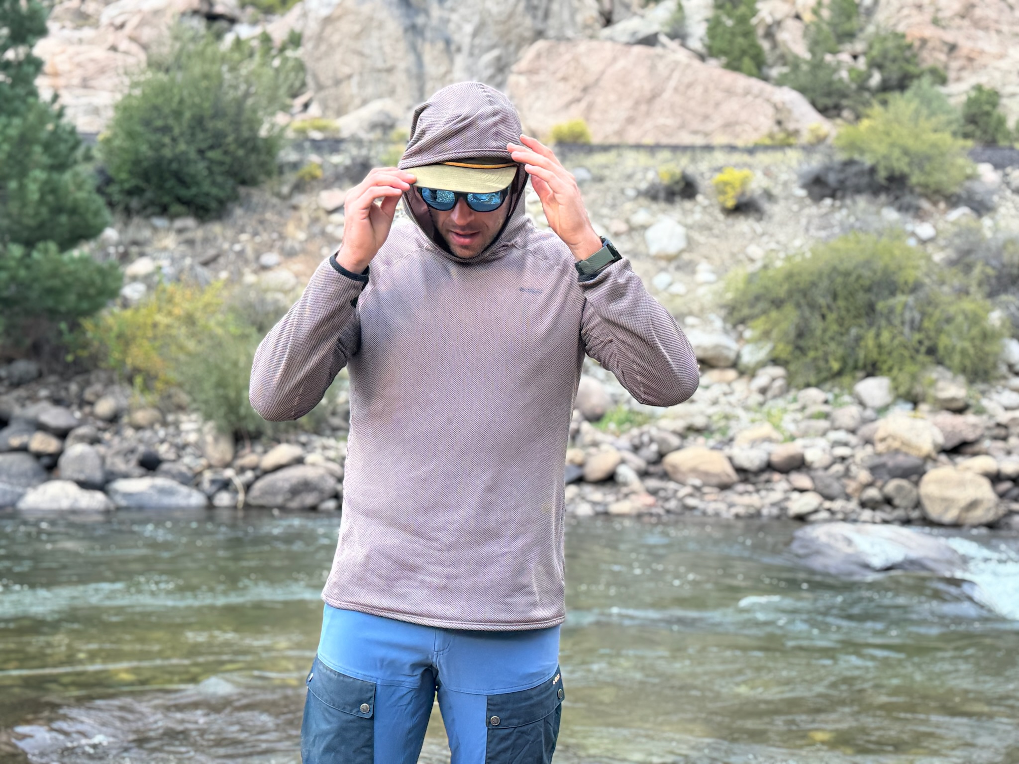 Man adjusts the hood of his brown hoody over a cap while standing by a river