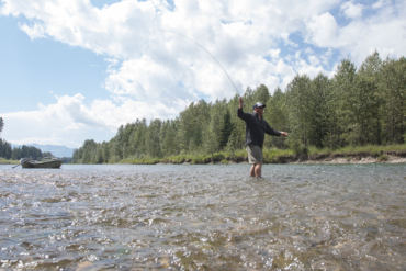 An angler casting a fly rod in a river with a driftboat in the background.