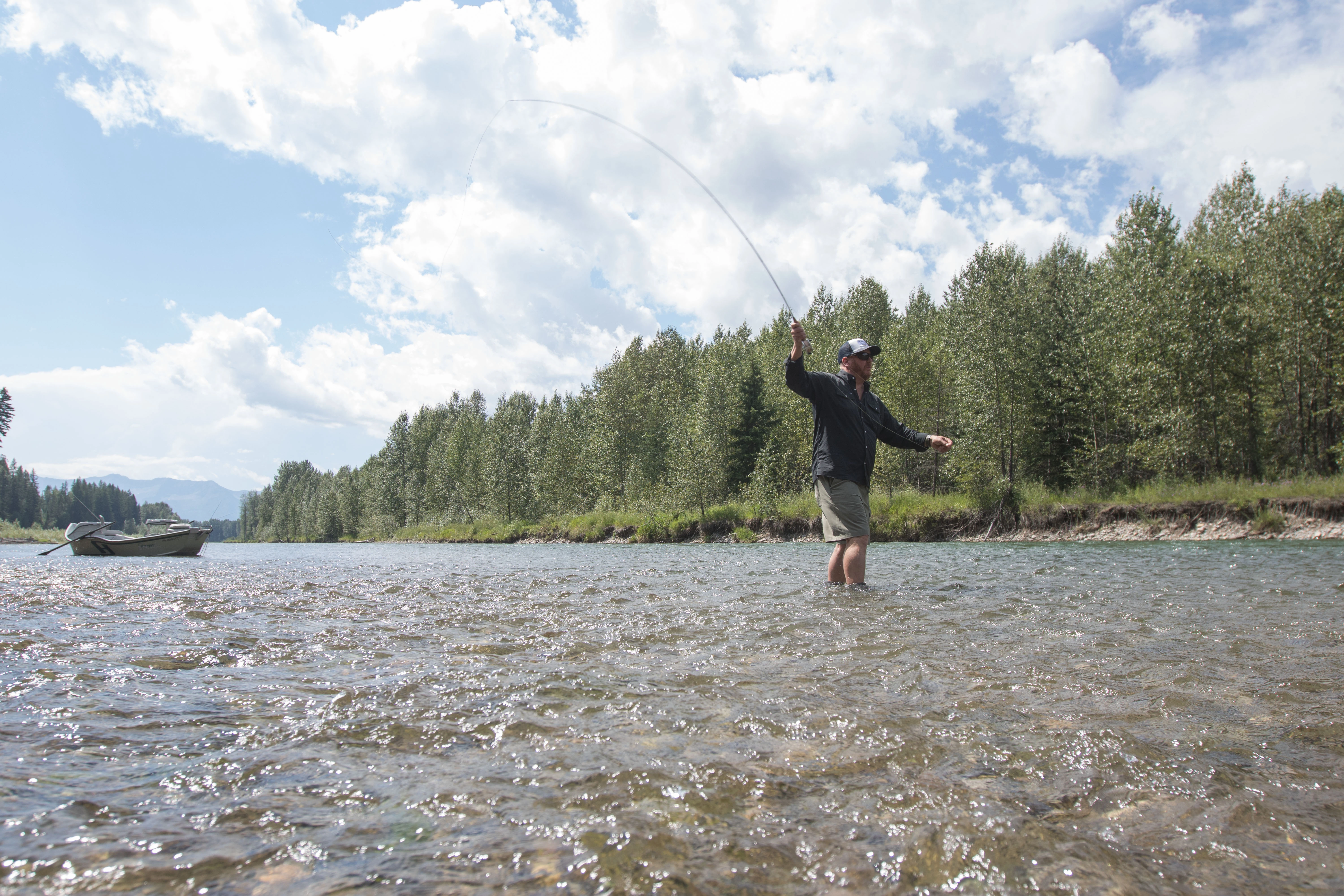 An angler casting a fly rod in a river with a driftboat in the background.