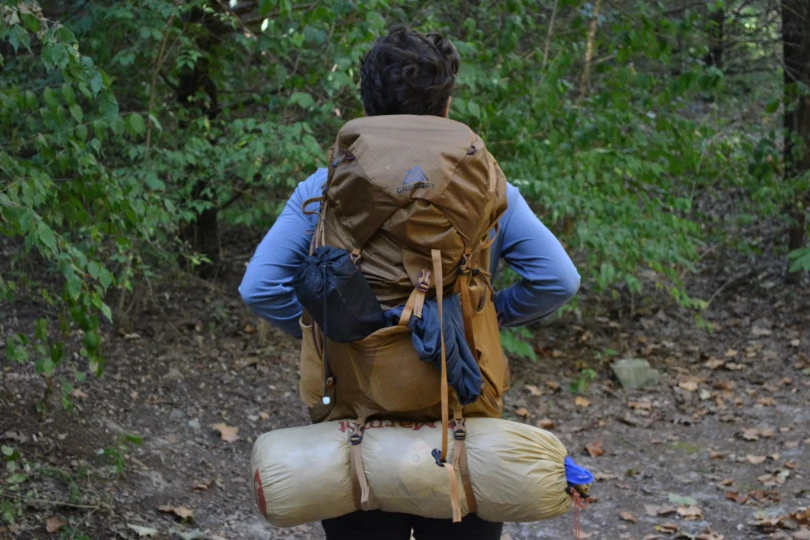 Person wearing brown Gregory backpack in woods, carrying tent and other tiems 