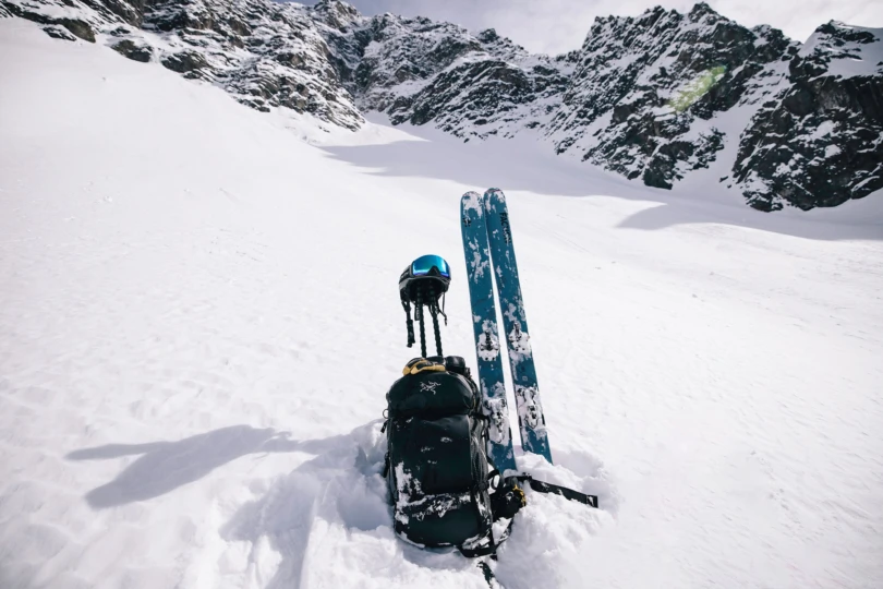 Blue skis standing in the snow with a black backpack, helmet, and poles beneath steep mountain walls