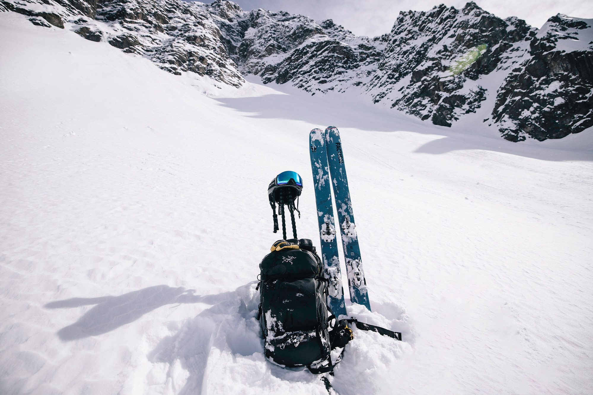 Blue skis standing in the snow with a black backpack, helmet, and poles beneath steep mountain walls