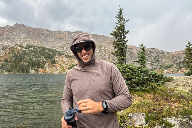 Man in the Mountain Hardwear Airmesh Hoody with hood up smiles while holding a water bottle near a lake