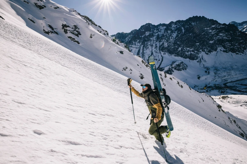 Skier hiking up a snowy slope in a yellow-brown jacket and green bibs under bright sun