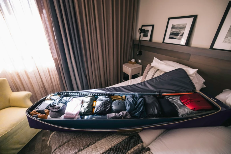 Large ski bag laid open on a hotel bed, neatly packed with gear, clothing, helmet, and poles