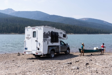A view of a truck camper and people carrying a canoe towards a lake in the mountains.