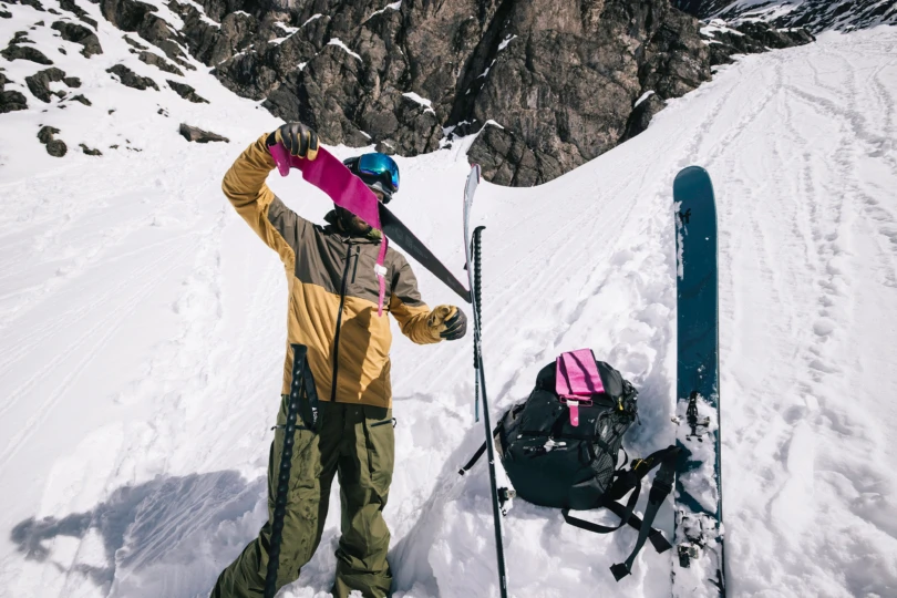 Skier adjusting bright pink climbing skins on blue skis while wearing a helmet and goggles