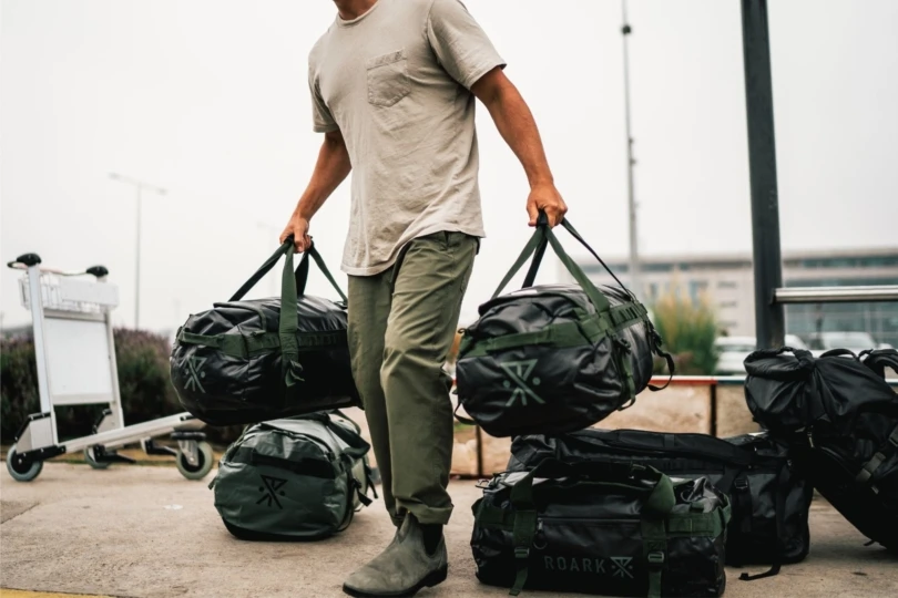 Person wearing white t-shirt and green pants loads duffel bags