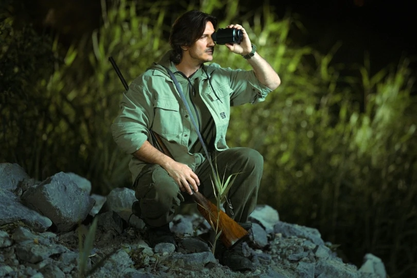 Person sits on rocky outcropping with hunting rifle and looking through a scope