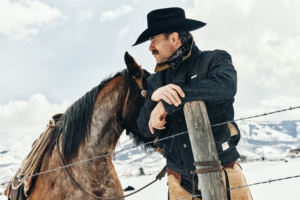 Cowboy leans against fence with horse in snowy landscape