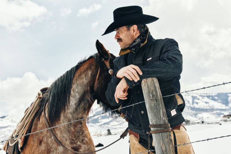 Cowboy leans against fence with horse in snowy landscape