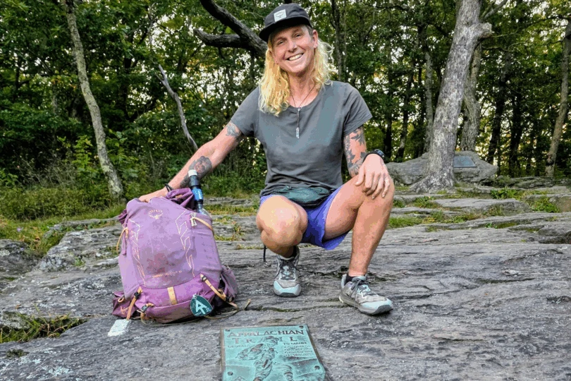 Woman with purple backpack kneels on rock next to AT sign. 