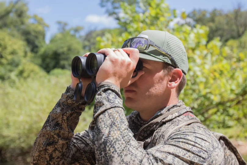 A hunter looking through binoculars with a forest in the background.