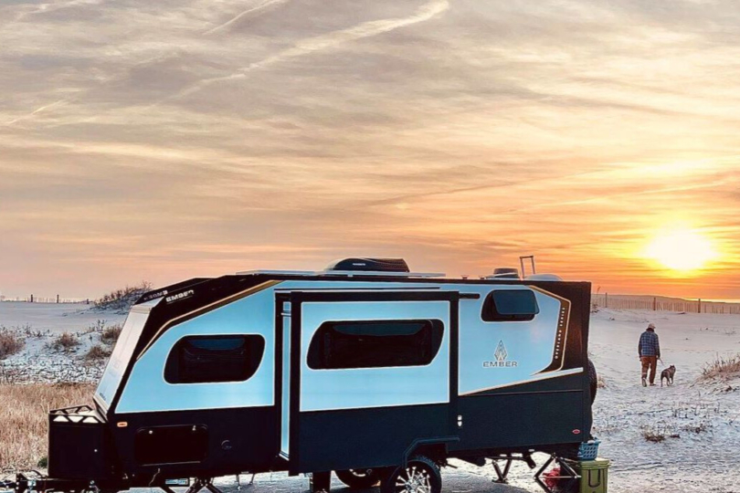 A person and their dog walks toward the sunset on a sand dune with a RV trailer parked in the foreground.