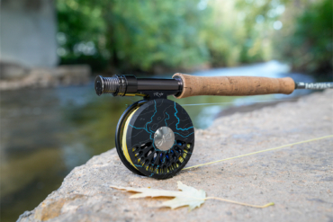 The custom reel rests on a riverside rock with soft green reflections in the background