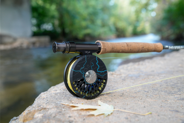 The custom reel rests on a riverside rock with soft green reflections in the background