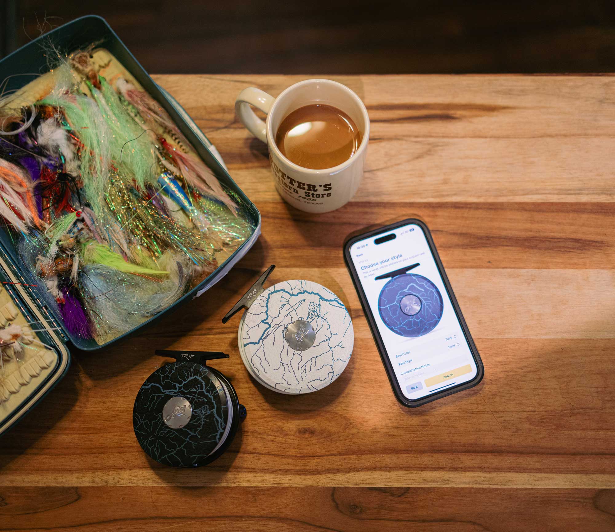 Two Abel x onWater Custom Reels shown on a wooden table beside a fly box, coffee mug, and a phone displaying reel customization options