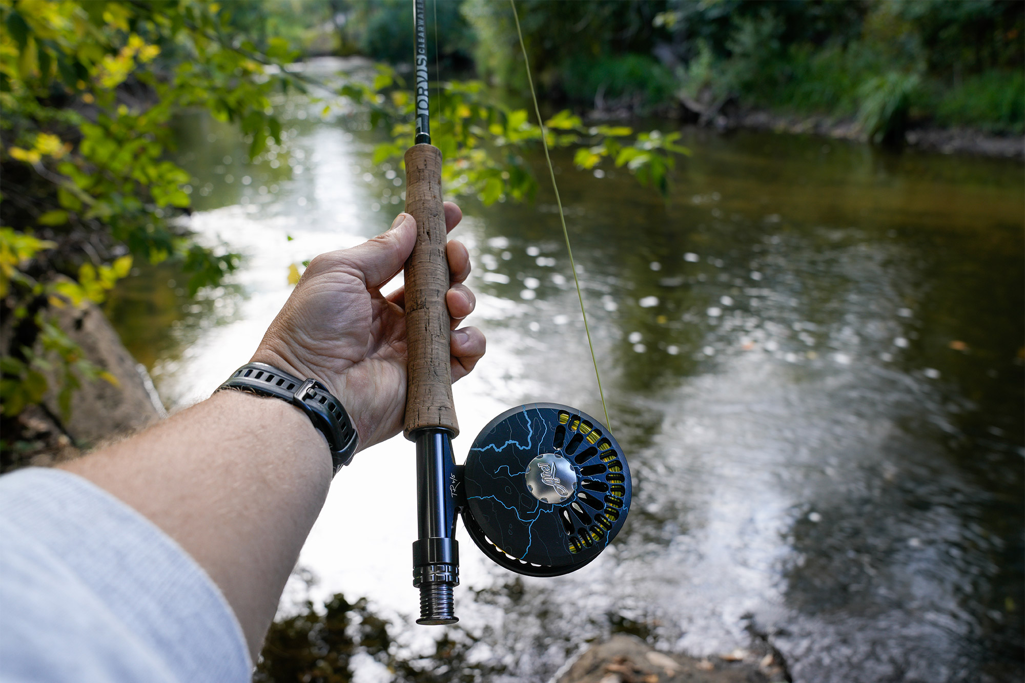 Angler holding a fly rod with the reel above the river, surrounded by green trees