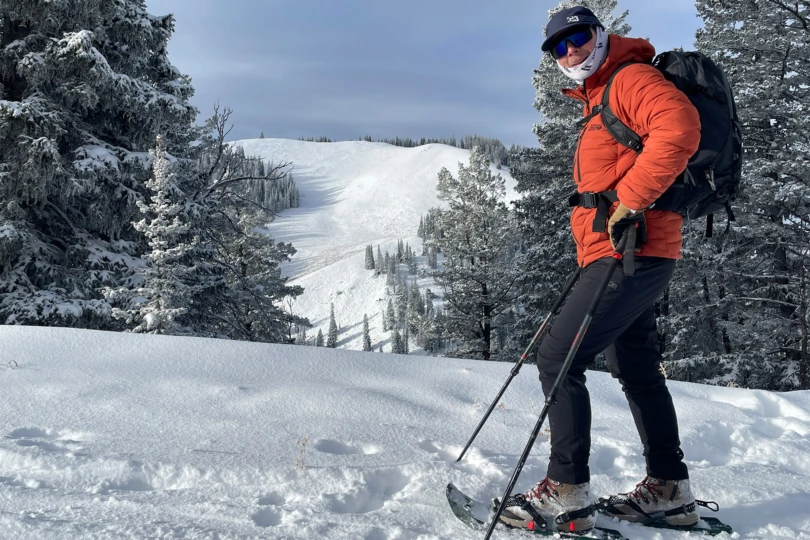 A hiker wearing Evo Trail snowshoes pauses on a ridge with snowy peaks ahead