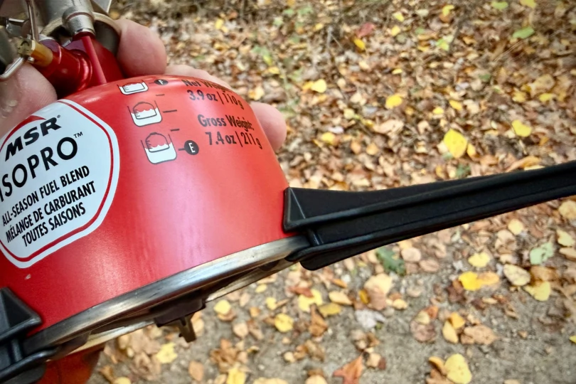 Close-up of the MSR Switch fuel canister showing the fill level markings and tripod stand outdoors on fallen leaves
