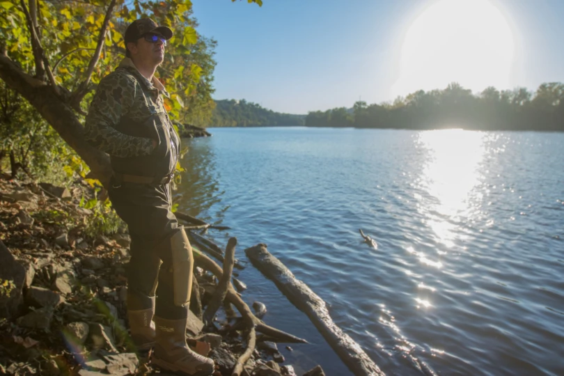A hunter on the side of a river wearing waders