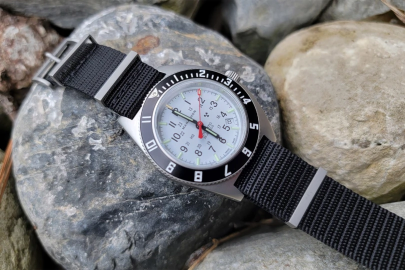 The Marathon SSNAV-D Auto Arctic Edition resting on smooth gray rocks, showing the contrast of its white dial and black strap